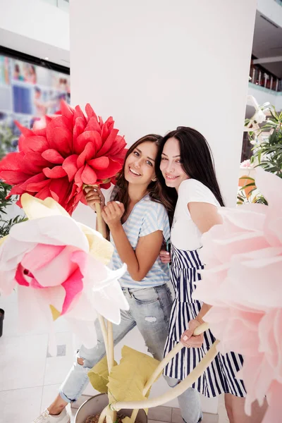 Dos chicas delgadas sonrientes y jóvenes, vestidas con ropa casual, se paran una al lado de la otra en un vestíbulo de un centro comercial moderno , — Foto de Stock