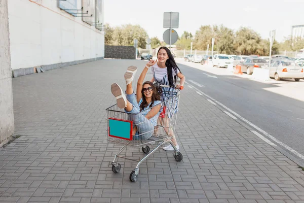 Deux jolies dames aux cheveux bruns, portant une tenue décontractée, s'amusent avec un chariot d'épicerie près du centre commercial . — Photo