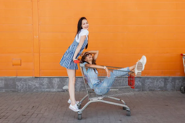 Duas senhoras muito magras de cabelos escuros, vestindo roupas casuais, se divertir com um carrinho de supermercado perto do centro comercial . — Fotografia de Stock