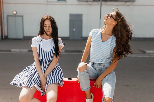 Dwie piękne dziewczyny slim z długimi włosami, noszenie stroju casual, siedź na płocie na drodze i uśmiech. — Zdjęcie stockowe