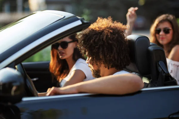 Jeune homme aux cheveux bruns Curly et deux belles filles aux cheveux bruns en lunettes de soleil sont assis dans un cabriolet noir . — Photo
