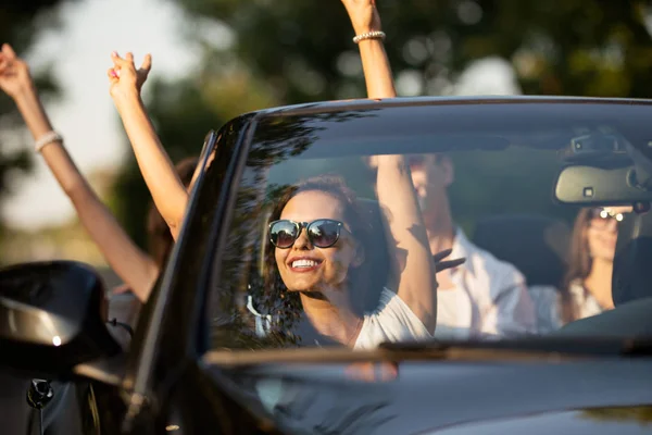 Kaunis brunette nuori nainen istuu ystävien kanssa musta cabriolet hymyilee ja pitää kätensä ylös aurinkoinen päivä . — kuvapankkivalokuva