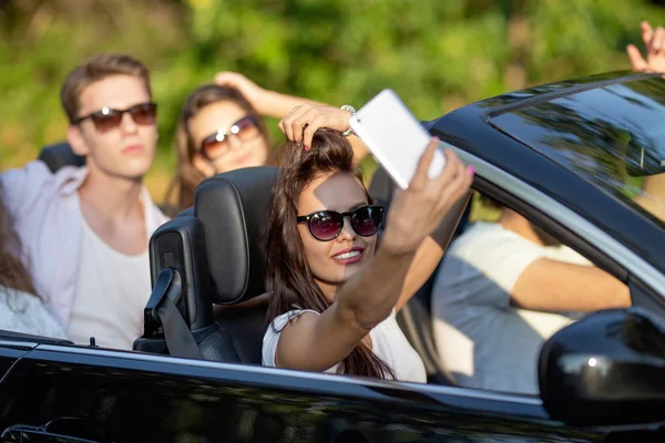 Attractive young brunette woman in sunglasses dressed in a white t-shirt sits with friends in a black cabriolet make a selfie on a sunny day. — Stock Photo, Image