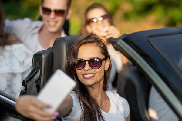 Mulher morena jovem atraente em óculos de sol vestida com uma camiseta branca senta-se com amigos em um cabriolet preto fazer uma selfie em um dia ensolarado . — Fotografia de Stock