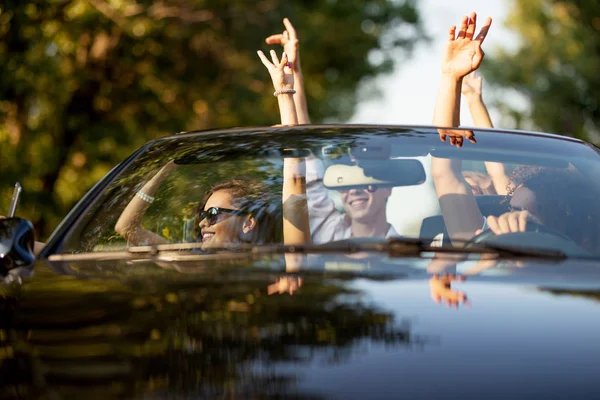 Young beautiful  peopple in sunglasses dressed in white clothes are sitting in a black cabriolet holding their hands up on a sunny day. — Stock Photo, Image