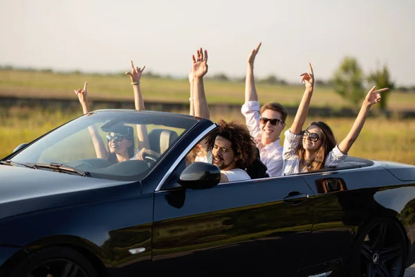 Belles jeunes filles et les gars en lunettes de soleil sourire et monter dans un cabriolet noir sur la route tenant leurs mains vers le haut par une journée ensoleillée . — Photo