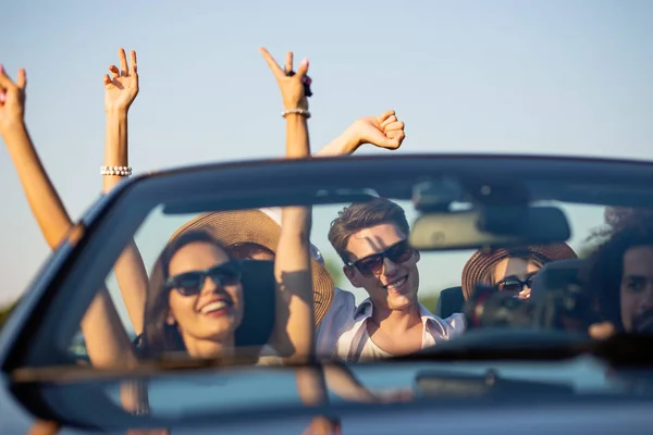 Meninas bonitas e caras de óculos de sol sorriem e andam em um cabriolet preto na estrada segurando as mãos em um dia ensolarado . — Fotografia de Stock