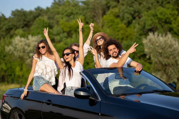 Lucky stylish young dark-haired girls and guys in sunglasses smile in a black cabriolet on the road holding their hands up on a sunny day. — Stock Photo, Image