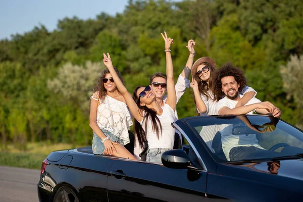 Cool stylish young dark-haired girls and guys in sunglasses smile in a black cabriolet on the road holding their hands up on a sunny day. — Stock Photo, Image