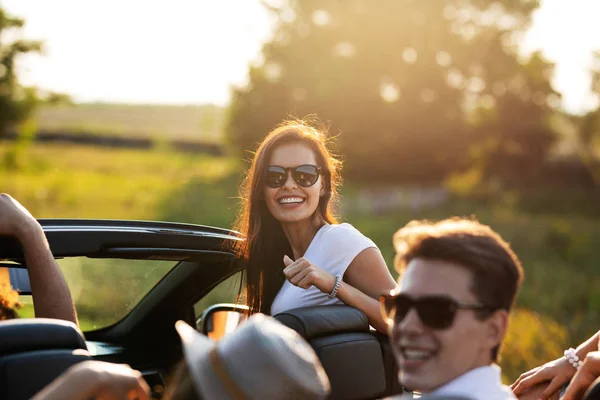 Linda jovem mulher de cabelos escuros em óculos de sol está sorrindo e sentado com os amigos em um cabriolet preto em um dia ensolarado . — Fotografia de Stock