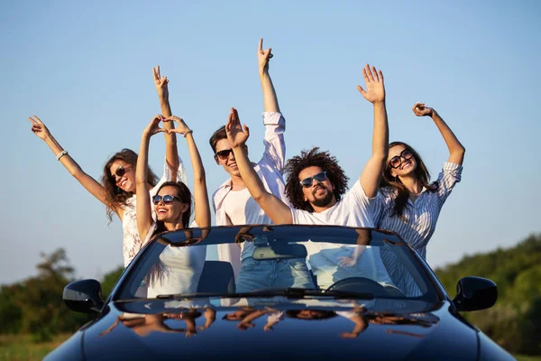 Happy young girls and guys in sunglasses are sitting in a black cabriolet on the road holding their hands up and smiling on a sunny day. — Stock Photo, Image