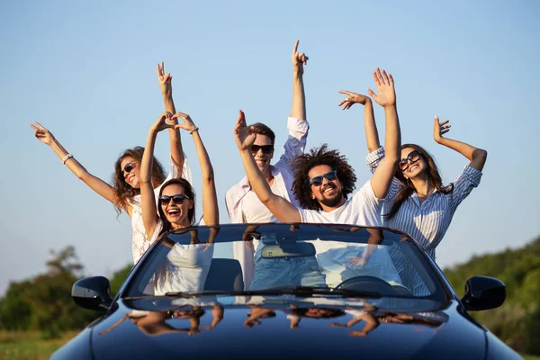 Happy young girls and guys in sunglasses are sitting in a black cabriolet on the road holding their hands up and smiling on a sunny day. — Stock Photo, Image