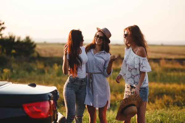 Drie mooie jonge meisjes in het veld naast de auto staan en praten op een warme zonnige dag. — Stockfoto