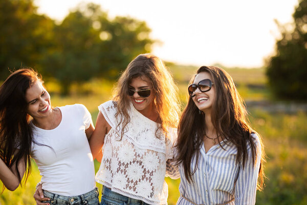 Three gorgeous dark-haired young women in sunglasses dressed in the beautiful clothes are smiling and walking outdoor on a sunny day.