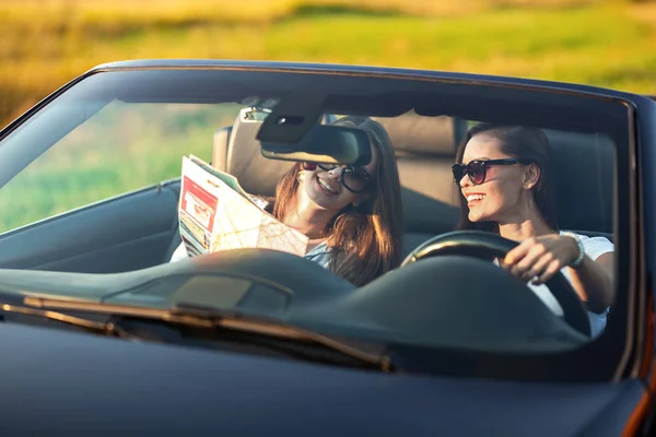 Kaksi tummahiuksista nuorta naista aurinkolaseissa istuvat mustassa cabrioletissa aurinkoisena päivänä. Yksi heistä pitää karttaa käsissään. . — kuvapankkivalokuva