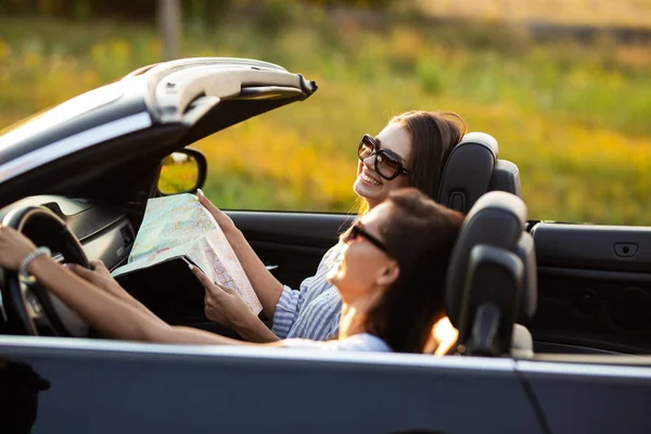 Two beautiful young women in sunglasses are sitting in a black cabriolet and smiling on a sunny day. One of them keeps map in her hands. — Stock Photo, Image