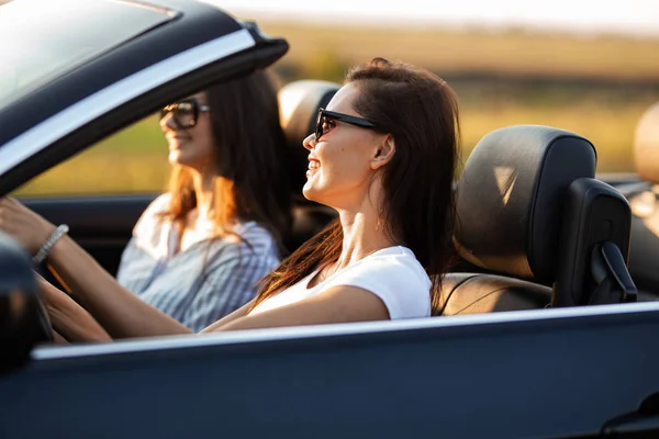 Deux belles jeunes femmes en lunettes de soleil sont assises dans un cabriolet noir et souriantes par une journée ensoleillée . — Photo