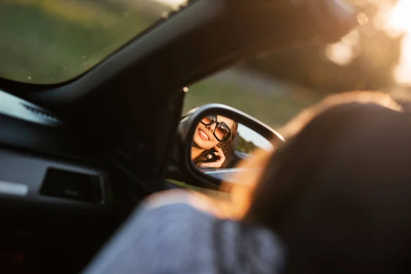 Reflexão de uma jovem menina sorridente de cabelos escuros em óculos de sol em um espelho lateral de um carro . — Fotografia de Stock