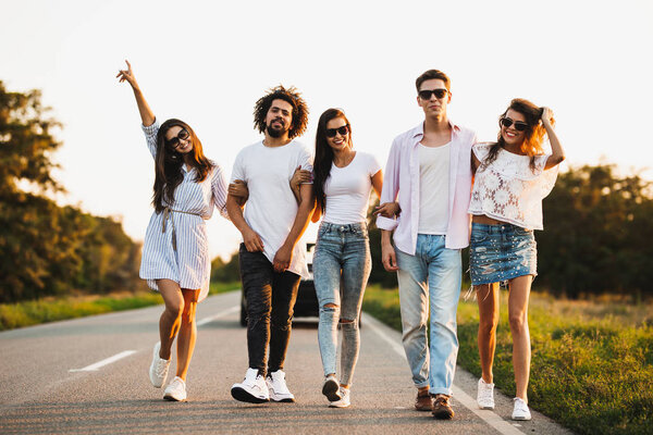 Young stylish two guys and three girls walk on a country road on a sunny day