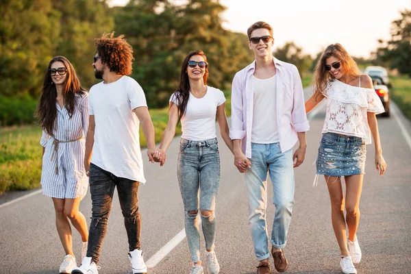 Jóvenes chicos y chicas felices están tomados de la mano y caminando en la carretera en un día soleado —  Fotos de Stock