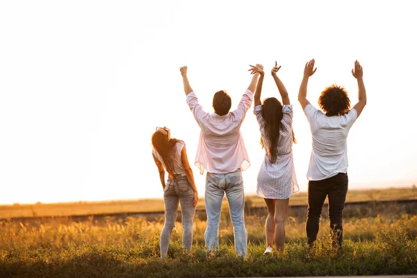 Vista posteriore. Due ragazzi e due ragazze sono in piedi sul campo in un giorno d'estate e tenendo le mani in alto . — Foto Stock
