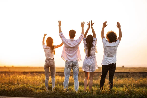 Vista posteriore. Compagnia di giovani ragazze e ragazzi sono in piedi sul campo in una giornata estiva e tenendo le mani in alto . — Foto Stock