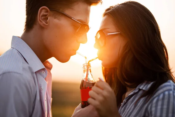 美丽的黑发女孩和一个戴着太阳镜的年轻人在阳光明媚的日子里, 从一个瓶子里喝了一杯, 在户外通过秸秆喝. — 图库照片