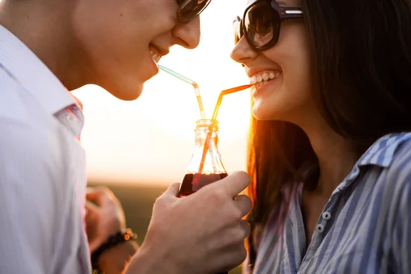 Beautiful dark-haired girl and a young man  in sunglasses are drinking from one bottle a drink through straws outdoor on a sunny day. — Stock Photo, Image