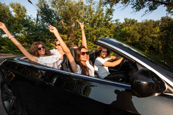 Compagnie de jeunes gens chevauchant dans un cabriolet sur la route par une chaude journée ensoleillée.. Deux belles filles tenant la main vers le haut . — Photo