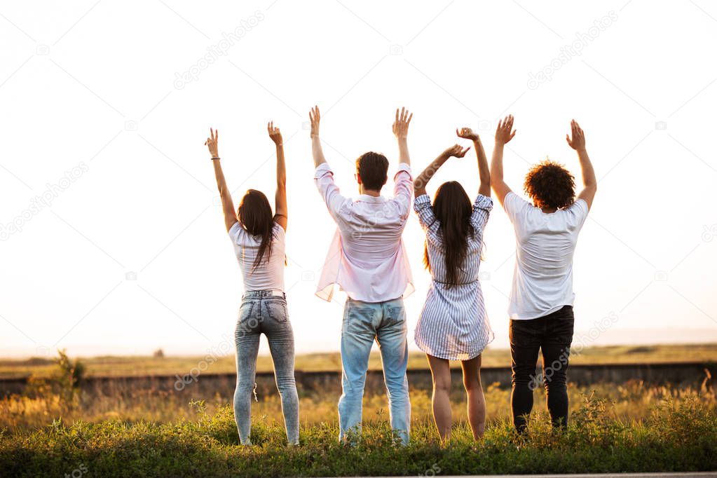Back view. Two guys and two girls are standing in the field on a summer day and holding their hands up.