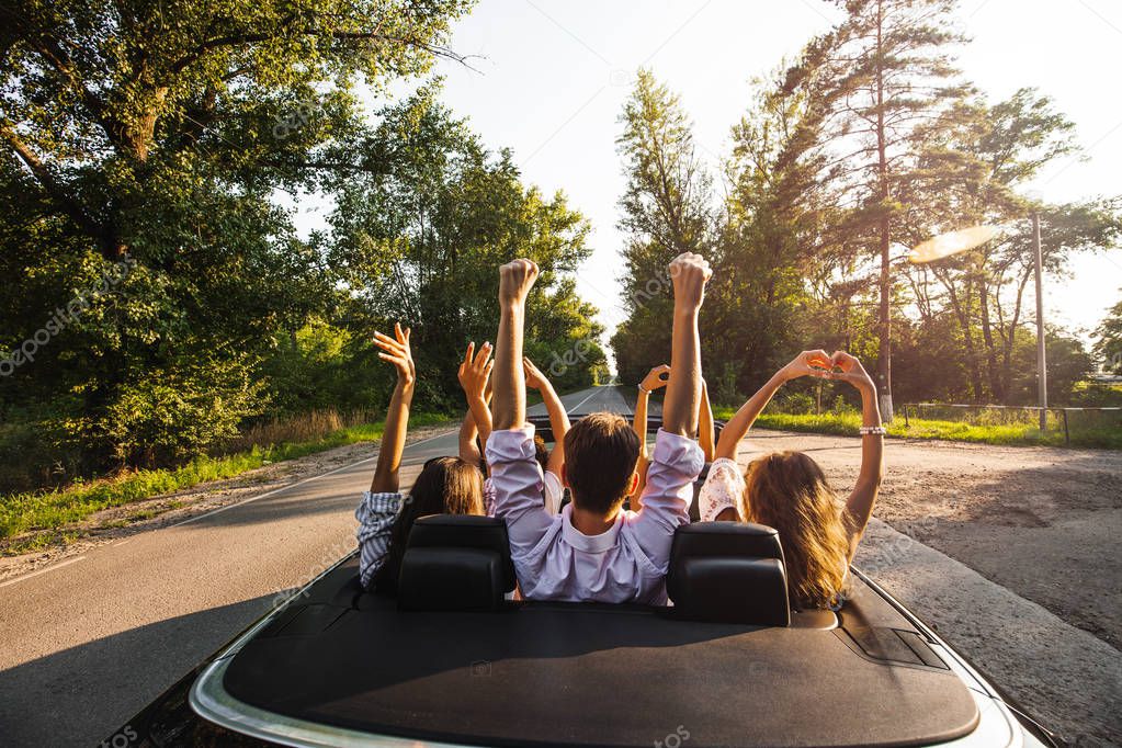 Company of young people riding in a cabriolet on the road and holding their hands up on a warm sunny day.