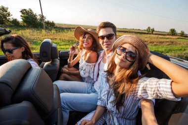 Şapkalar iki genç esmer kadın genç adamla bir siyah cabriolet oturan ve bir yaz gününde gülümseyen.