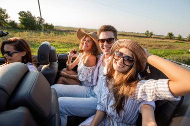Şapkalar iki genç esmer kadın genç adamla bir siyah cabriolet oturan ve bir yaz gününde gülümseyen.