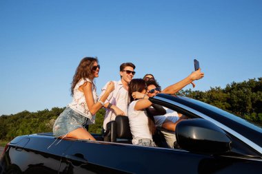 Neşeli genç kızlar ve çocuklar yolda siyah bir Cabriolet oturma ve selfie güneşli bir günde yapma.