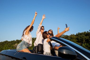 Neşeli genç kızlar ve çocuklar yolda siyah bir Cabriolet oturma ve selfie güneşli bir günde yapma.