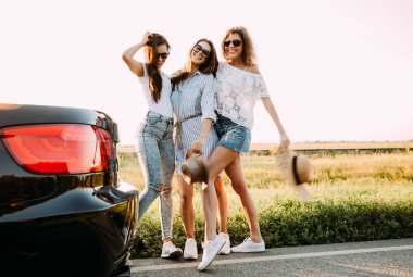 Üç güzel genç kız arabanın yanındaki alanda stand ve sıcak güneşli bir günde konuşmak.