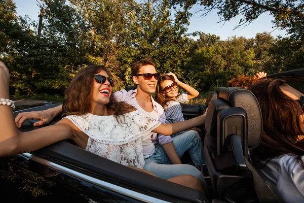 Company of young people riding in a cabriolet on the road on a warm sunny day. Two beautiful girls and a young man between them seats on the backseat and smile. — Stock Photo, Image
