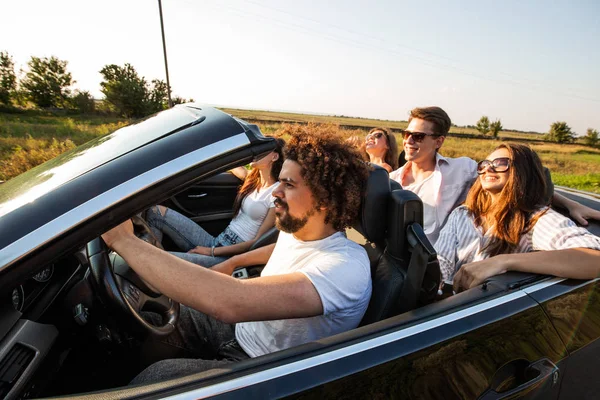 Group of young beautiful girls and guys in sunglasses   smile and ride in a black cabriolet on the road on a sunny day. — Stock Photo, Image