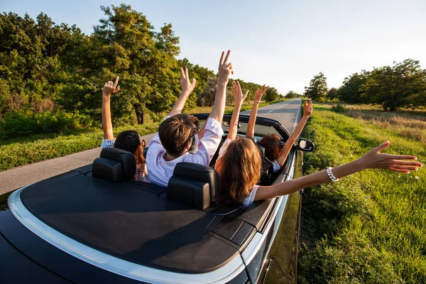 Company of young girls and guys are sitting in a black cabriolet hold their hands up on the country road on a sunny day. — Stock Photo, Image