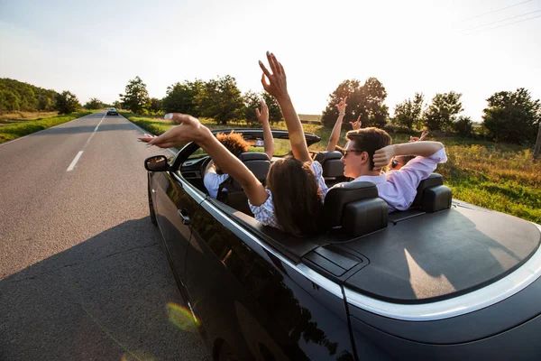 O cabriolet preto está na estrada do campo. Companhia de meninas e caras estão sentados no carro segurar as mãos para cima em um dia ensolarado . — Fotografia de Stock