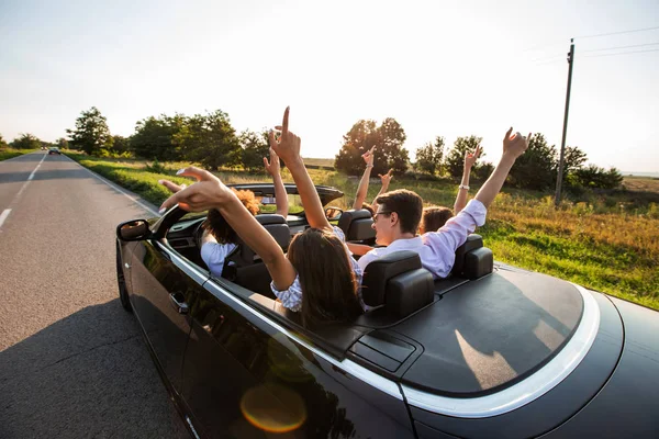 Le cabriolet noir est sur la route de campagne. Heureux groupe de jeunes filles et les gars sont assis dans la voiture tenir leurs mains vers le haut par une journée ensoleillée . — Photo