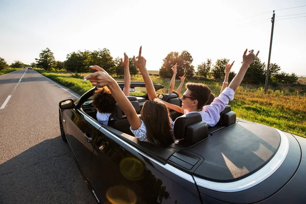 O cabriolet preto está na estrada do campo. grupo feliz de meninas e caras estão sentados no carro segurar as mãos para cima em um dia ensolarado . — Fotografia de Stock