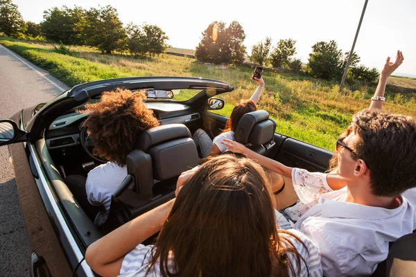 Les jeunes filles et les gars sont assis dans une voiture décapotable noire et faire la photo au téléphone au coucher du soleil — Photo