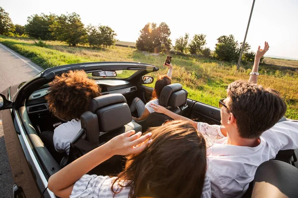 Young girls and guys are sitting in a black cabriolet and making selfie on a warm sunny day. — Stock Photo, Image