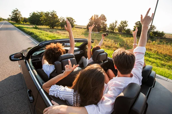 Jeunes filles et les gars sont assis dans un cabriolet noir, tenant leurs mains vers le haut et faire du selfie par une chaude journée ensoleillée . — Photo