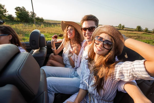 Duas jovens mulheres de cabelos escuros em chapéus estão sentadas jovens com o homem em um cabriolet preto e sorrindo em um dia de verão. . — Fotografia de Stock