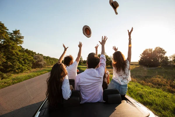 Company of happy young girls and guys sit in a black cabriolet road and throw up their hats on a sunny day.