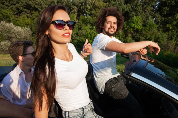 Wunderschöne junge dunkelhaarige Frau mit Sonnenbrille und junger Mann sitzen in einem schwarzen Cabriolet an einem sonnigen Tag. — Stockfoto