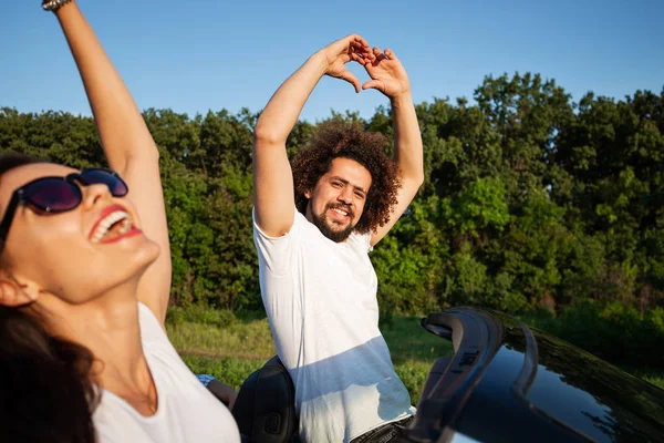 Curly jovem de cabelos escuros segurando as mãos para cima, sentado com uma bela jovem mulher em um cabriolet preto em um dia ensolarado . — Fotografia de Stock