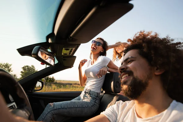 Curly dark-haired young man sitting at the wheel of a black cabriolet and young woman is sitting next to him on a sunny day. — Stock Photo, Image